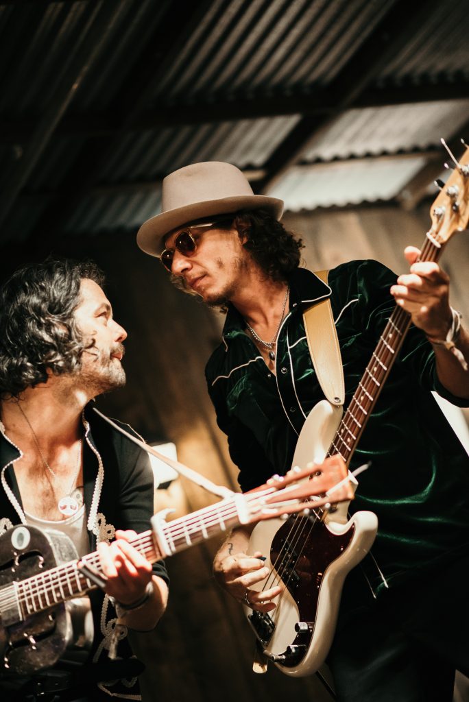Close-up of the duo mid-song, one on a resonator guitar, the other on bass in a velvet shirt and wide-brimmed hat