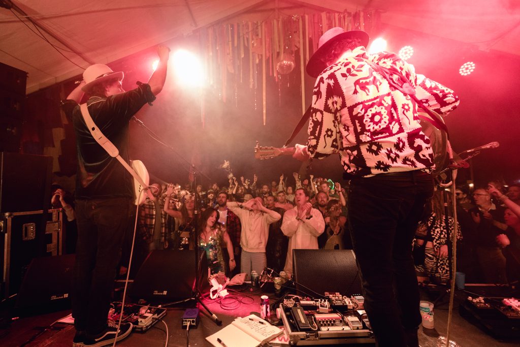 Two of the band in wide-brimmed hats facing each other on a festival tent stage lit deep red, the front row singing along