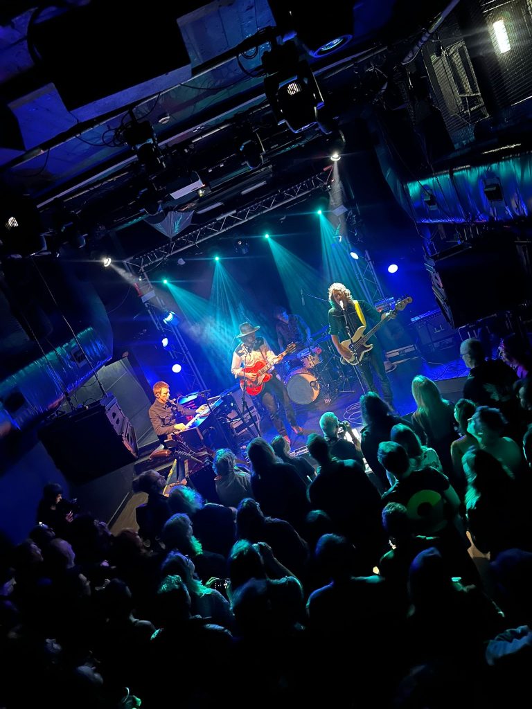 True Strays on stage in blue and green light at Thekla in Bristol, seen over the heads of a packed crowd